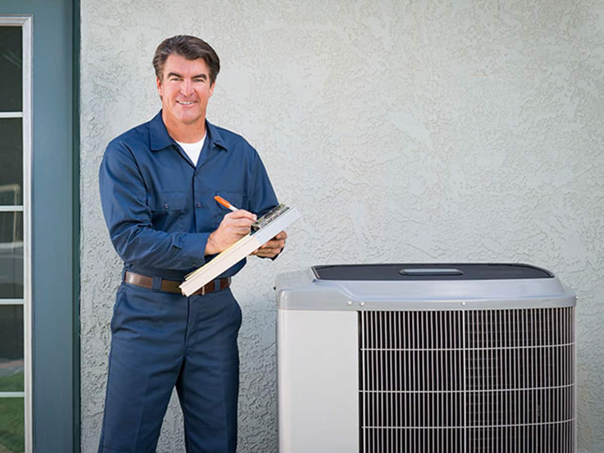 A man in blue work clothes standing next to an air conditioner.